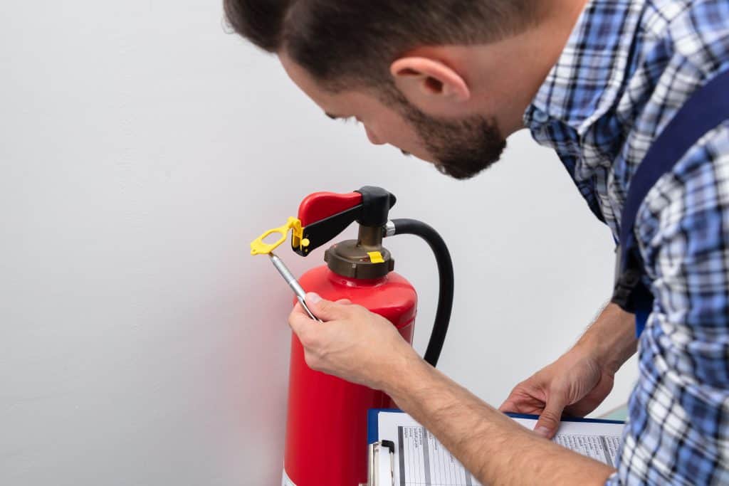 A person in a plaid shirt inspects a red fire extinguisher, holding a pen and clipboard, possibly checking pressure or recording maintenance details—an example of NY Fire Protection Services Long Island ensuring safety compliance.