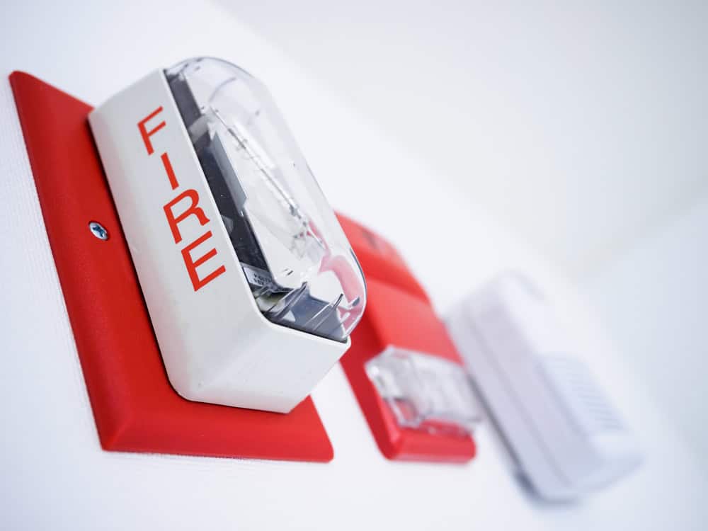 Close-up photograph of a wall-mounted fire alarm system. The focal point is a white and red alarm box with the word "FIRE" written on it in bold red letters. Additional components, essential for fire extinguisher installation on Long Island, are visible in the background, slightly out of focus.