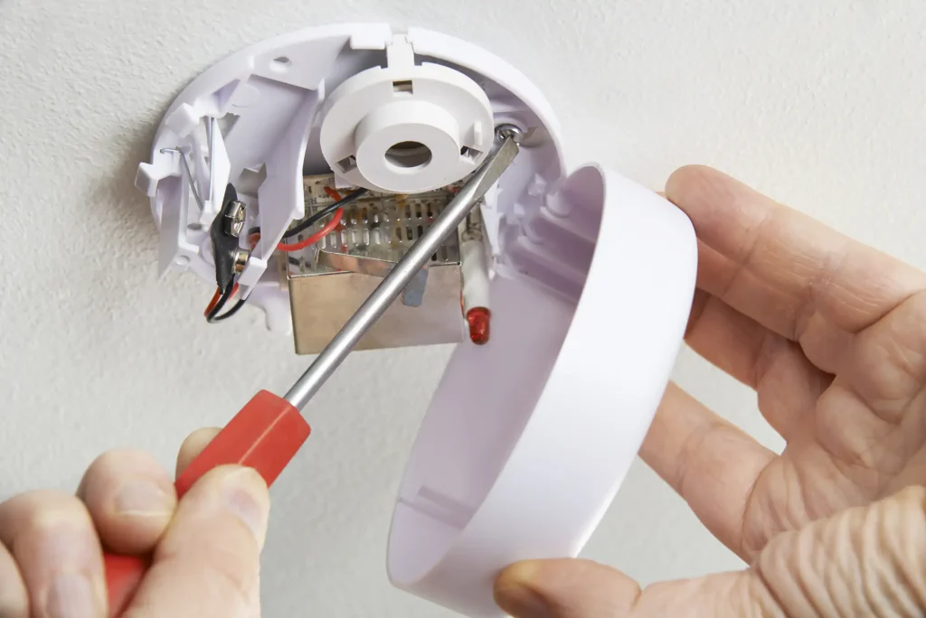 A person in NY uses a screwdriver to install or repair a smoke detector on a ceiling, holding the device’s cover and exposing its internal wires and components—a key step in Fire Protection Services Long Island professionals provide.