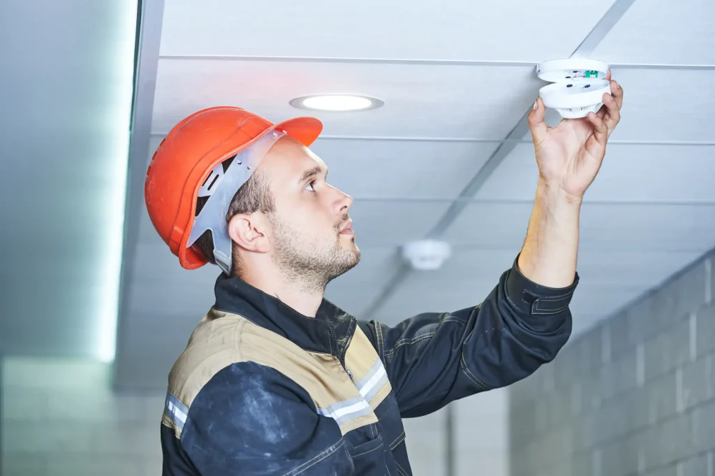 A worker wearing an orange hard hat and work jacket installs or checks a smoke detector on a ceiling in a modern NY building, ensuring top-notch Fire Protection Services Long Island can rely on.