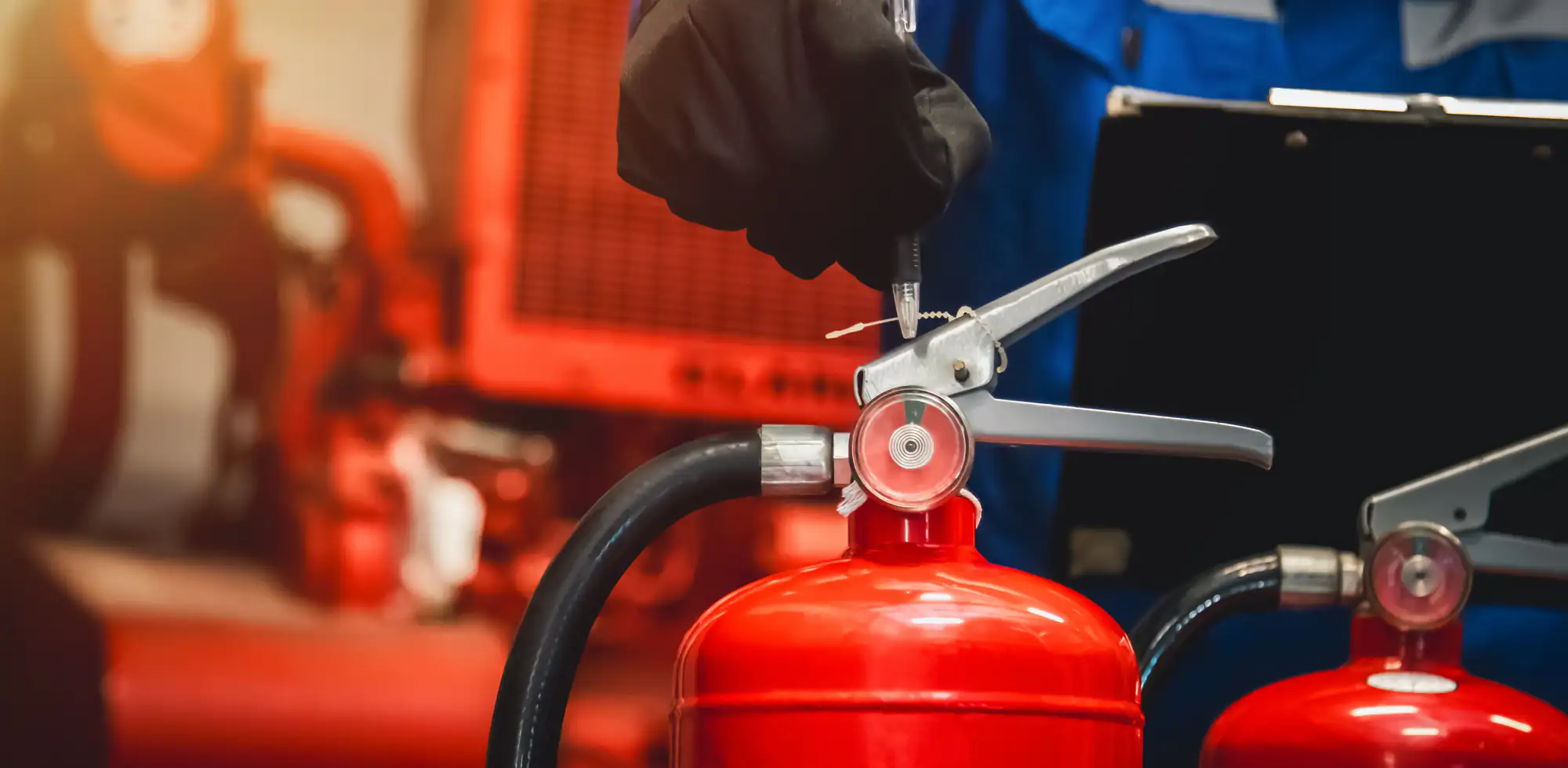 A person wearing a black glove inspects a red fire extinguisher, highlighting the importance of Fire Protection Services Long Island, NY, with industrial equipment and another extinguisher in the blurred background.