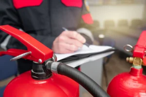 A person in a navy and red uniform writes on a clipboard in the background, while two red fire extinguishers—part of Fire Protection Services Long Island, NY—are in focus in the foreground.
