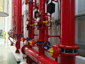 Rows of red industrial fire protection piping and valves, mounted vertically and horizontally, inside a building with a concrete floor and metal walls, installed by Fire Protection Services Long Island, NY.