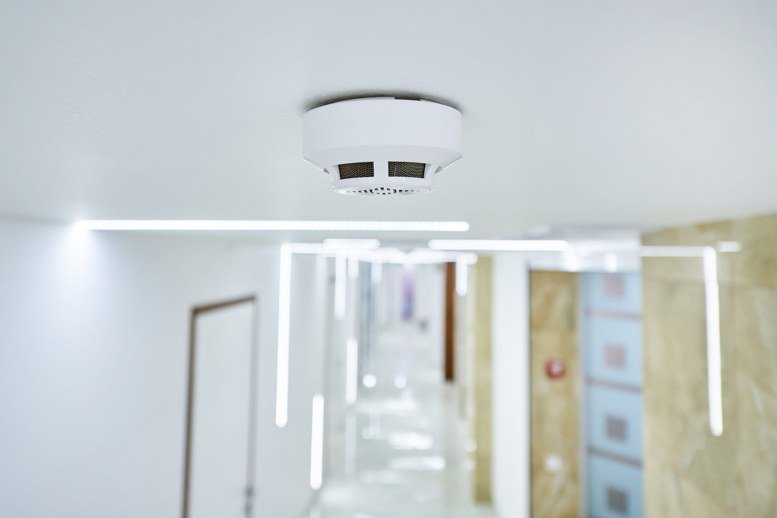 A smoke detector is mounted on a white ceiling in a brightly lit modern hallway, reflecting the commitment to fire protection services in Long Island, NY, with white walls, doors, and decorative lighting strips.