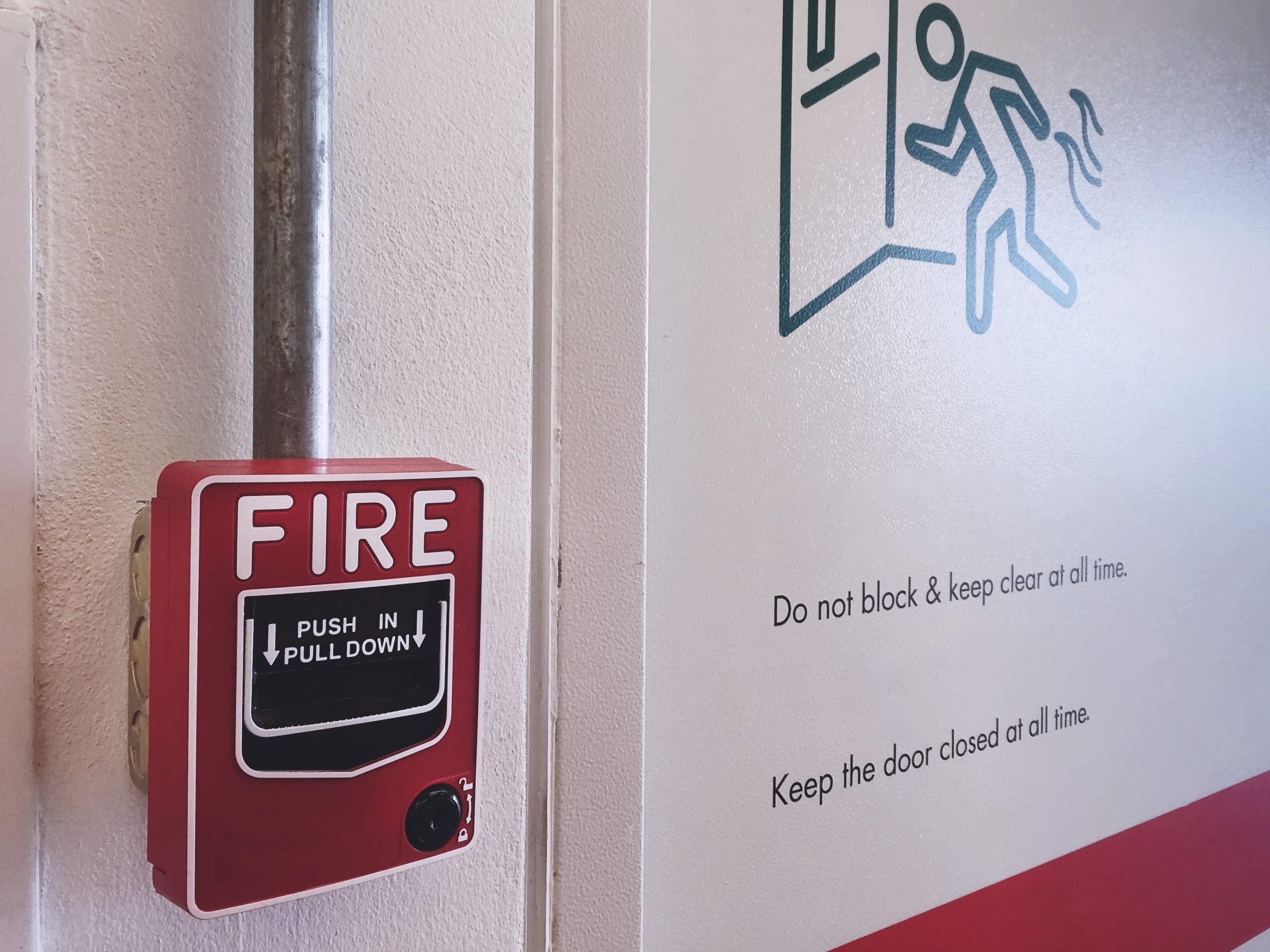 A red fire alarm box labeled “FIRE” is mounted on a wall near a metal pipe. In this NY building, Fire Protection Services Long Island ensures safety—signs remind: “Do not block & keep clear at all time. Keep the door closed at all time.”.