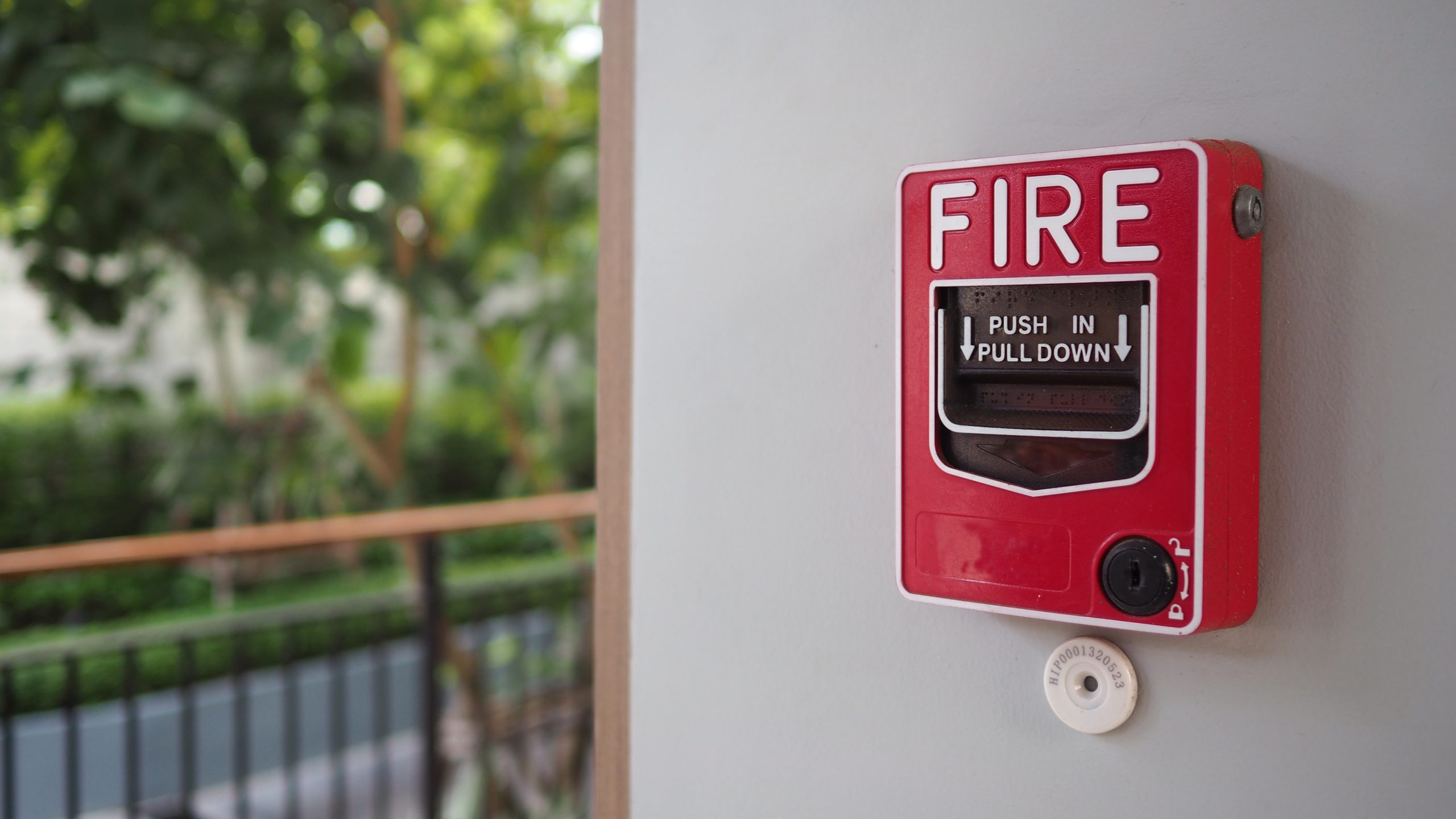 A red fire alarm pull station mounted on a light gray wall serves as a crucial safety feature, reflecting the reliability of Fire Protection Services Long Island, NY, with greenery and outdoor railings blurred in the background.