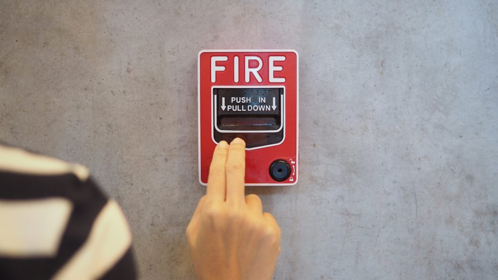 A person’s hand reaches to pull down a red fire alarm on a gray wall, following the "PUSH IN PULL DOWN" instructions. Part of their striped sleeve is visible—a reminder of the importance of Fire Protection Services Long Island, NY.