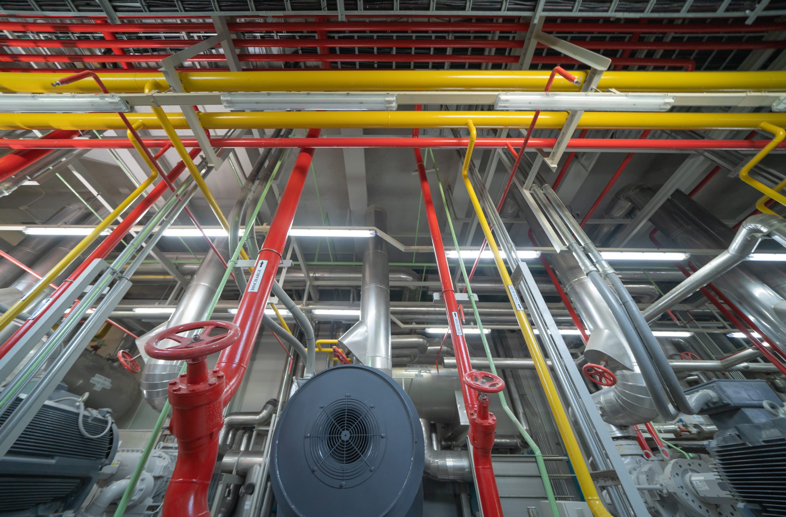 Large industrial pipes and machinery with red, yellow, and silver pipes running across the ceiling, featuring valves and complex metal structures in a brightly lit NY facility, viewed from a low angle. Fire Protection Services Long Island compliant.
