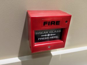 A red fire alarm box mounted on a beige wall in NY, with instructions that say "BREAK GLASS PRESS HERE" beneath a "FIRE" label—an essential feature for Fire Protection Services Long Island.