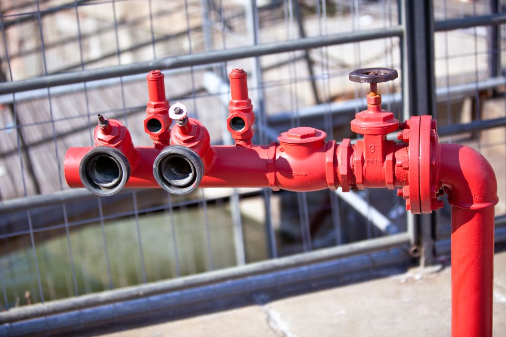A close-up of a red fire hydrant with multiple valves and two hose connections, situated outdoors near a metal fence—ideal for Fire Protection Services Long Island, NY.